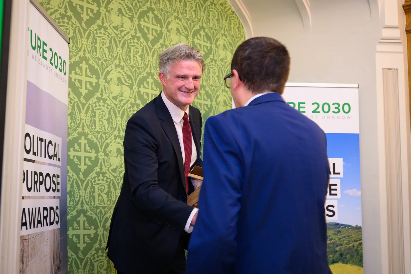 A man in a dark suit hands a wooden trophy to another man in a navy suit, in front of Political Purpose Awards and Nature 2030 banners